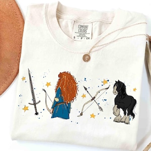 May include: Cream-colored t-shirt featuring a cartoon illustration of a red-haired character in a blue dress, a sword, a bow and arrow, a black horse, and yellow stars. The shirt has a Comfort Colors tag.