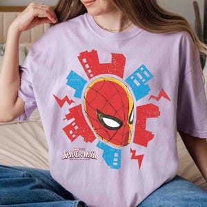 Puede incluir: Una camiseta de manga corta de color morado claro con un gr&aacute;fico de la m&aacute;scara de Spider-Man rodeada de siluetas de edificios rojos y azules y rayos. El texto Spider-Man Brand New Day est&aacute; impreso en blanco debajo de la m&aacute;scara.