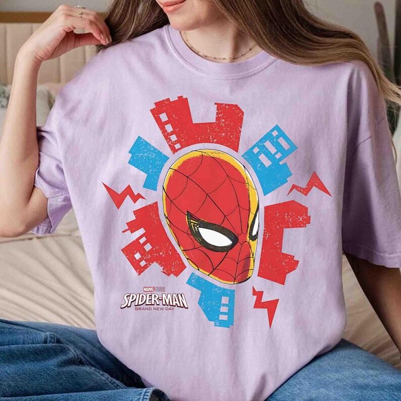 Puede incluir: Una camiseta de manga corta de color morado claro con un gr&aacute;fico de la m&aacute;scara de Spider-Man rodeada de siluetas de edificios rojos y azules y rayos. El texto Spider-Man Brand New Day est&aacute; impreso en blanco debajo de la m&aacute;scara.