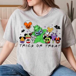 May include: A light gray t-shirt featuring a Halloween-themed graphic. The design includes cartoon characters, balloons, and the text "TRICK OR TREAT" in black. The shirt is a casual, short-sleeved style.