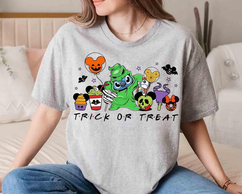 May include: A light gray t-shirt featuring a Halloween-themed graphic. The design includes cartoon characters, balloons, and the text "TRICK OR TREAT" in black. The shirt is a casual, short-sleeved style.