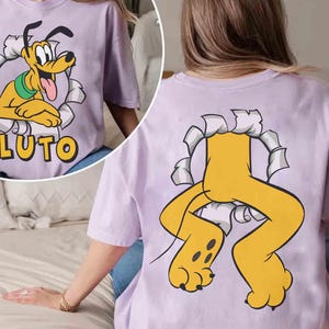 May include: A lilac t-shirt with a cartoon image of Pluto, the Disney dog, breaking through a paper design. The word "PLUTO" is printed in yellow. The back of the shirt shows Pluto's legs and paws.