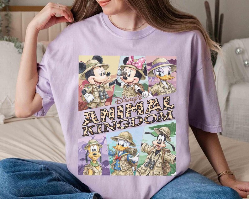 K&ouml;nnte beinhalten: Helllila T-Shirt mit einem Aufdruck von Disney-Figuren Mickey, Minnie, Donald, Goofy und Daisy in Safari-Outfits. Der Text "Disney's Animal Kingdom" ist unter den Figuren aufgedruckt.