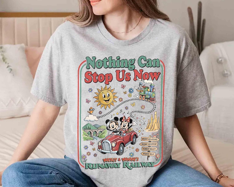 Peut inclure: Un t-shirt gris avec un motif color&eacute; affichant le texte "Nothing Can Stop Us Now". Le motif comprend Mickey et Minnie Mouse dans une voiture rouge, un soleil souriant et un train. Le t-shirt a un col rond et des manches courtes.