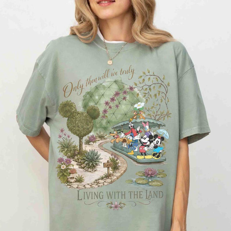May include: Sage green t-shirt featuring a whimsical illustration of Disney characters on a boat ride. The design includes the text "Only then will we truly" and "Living with the Land". The shirt has a relaxed fit.
