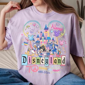 May include: A light purple t-shirt featuring a colorful Disneyland graphic. The design includes the Disneyland logo, various Disney characters, a castle, and the text "70 Years of Magic 1955-2025". The shirt has short sleeves.