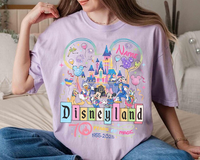 May include: A light purple t-shirt featuring a colorful Disneyland graphic. The design includes the Disneyland logo, various Disney characters, a castle, and the text "70 Years of Magic 1955-2025". The shirt has short sleeves.