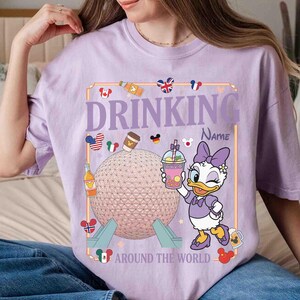 Puede incluir: Camiseta de color lila claro con un dise&ntilde;o gr&aacute;fico. El dise&ntilde;o incluye a Daisy Duck sosteniendo una bebida, el texto "DRINKING" encima de un campo de nombre y "AROUND THE WORLD" debajo. El gr&aacute;fico tambi&eacute;n presenta un globo, banderas e iconos de bebidas.