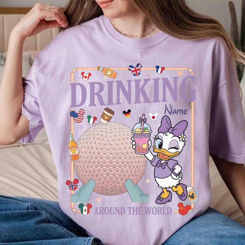 Puede incluir: Camiseta de color lila claro con un dise&ntilde;o gr&aacute;fico. El dise&ntilde;o incluye a Daisy Duck sosteniendo una bebida, el texto "DRINKING" encima de un campo de nombre y "AROUND THE WORLD" debajo. El gr&aacute;fico tambi&eacute;n presenta un globo, banderas e iconos de bebidas.