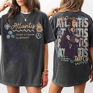 Könnte beinhalten: Dunkelgraues T-Shirt mit dem Wort "Atlantis" und nautischen Grafiken. Auf der Vorderseite steht "Chart a Course to Atlantis". Auf der Rückseite befindet sich eine Cartoon-Illustration eines Charakters und die Worte "Milo" und "Thatch".
