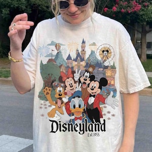 May include: Cream-colored t-shirt featuring a colorful Disneyland graphic with Mickey Mouse, Minnie Mouse, Goofy, Donald Duck, and Pluto. The graphic includes a castle, fireworks, and a Ferris wheel. The text "Disneyland Est. 1955" is printed below the characters.