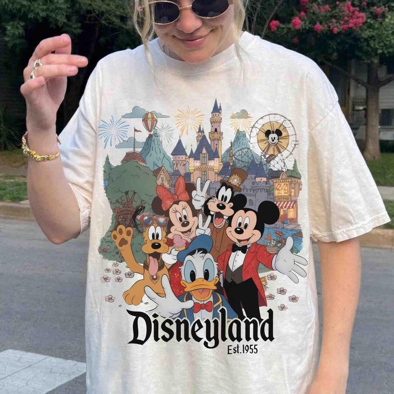 May include: Cream-colored t-shirt featuring a colorful Disneyland graphic with Mickey Mouse, Minnie Mouse, Goofy, Donald Duck, and Pluto. The graphic includes a castle, fireworks, and a Ferris wheel. The text "Disneyland Est. 1955" is printed below the characters.