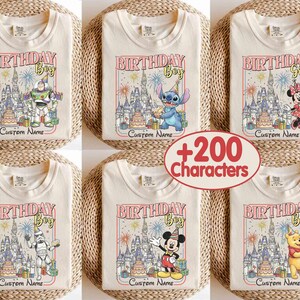 May include: Cream-colored t-shirts with colorful graphic designs featuring Disney characters and the text "BIRTHDAY Boy" or "BIRTHDAY Girl". Each shirt has a castle illustration, fireworks, and space for a custom name. The shirts are folded on woven mats.