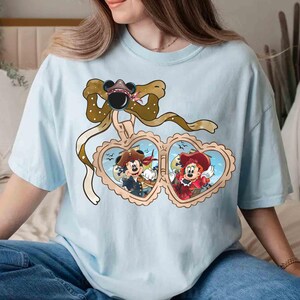 May include: Light blue t-shirt with a brown bow and two heart-shaped frames. The frames contain illustrations of Mickey Mouse and Minnie Mouse dressed as pirates.