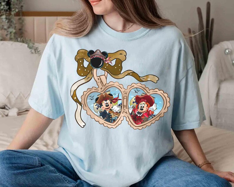 May include: Light blue t-shirt with a brown bow and two heart-shaped frames. The frames contain illustrations of Mickey Mouse and Minnie Mouse dressed as pirates.
