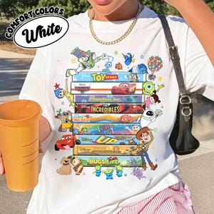 May include: White t-shirt featuring a stack of colorful movie titles and characters from Disney and Pixar films, including "Toy Story," "Cars," and "Monsters, Inc." The shirt has the words "COMFORT COLORS" and "White" printed on it.