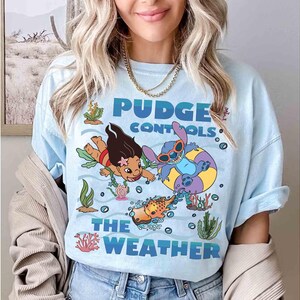 May include: Light blue t-shirt featuring a graphic of Lilo and Stitch in an underwater scene. The text "Pudge Controls The Weather" is displayed in blue. The design includes cartoon characters, coral, and seaweed.