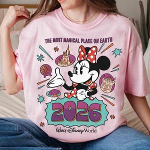 May include: Pale pink t-shirt with a Minnie Mouse graphic, wearing a red polka dot bow, pointing towards the text "2026" and "Walt Disney World". The shirt also features the text "The Most Magical Place on Earth" and Disney illustrations.