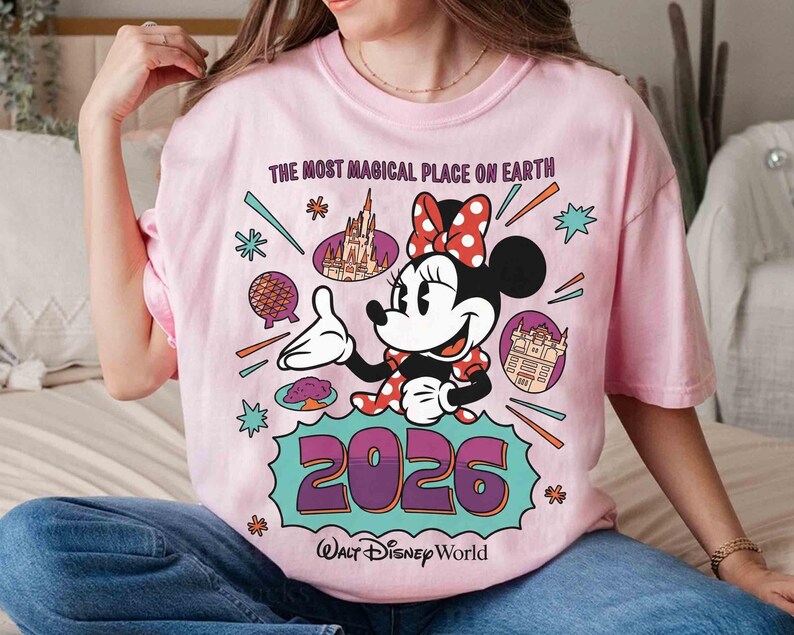 May include: Pale pink t-shirt with a Minnie Mouse graphic, wearing a red polka dot bow, pointing towards the text "2026" and "Walt Disney World". The shirt also features the text "The Most Magical Place on Earth" and Disney illustrations.