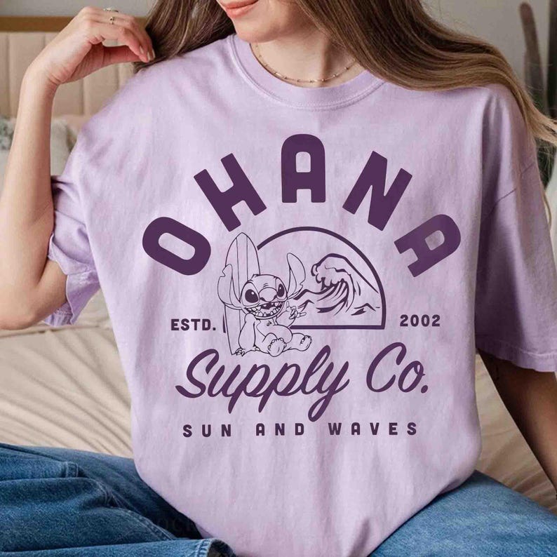 May include: A light purple t-shirt with a graphic featuring the word "OHANA" above a cartoon character holding a surfboard, with "Supply Co." and "Sun and Waves" below. The shirt also has "ESTD. 2002".
