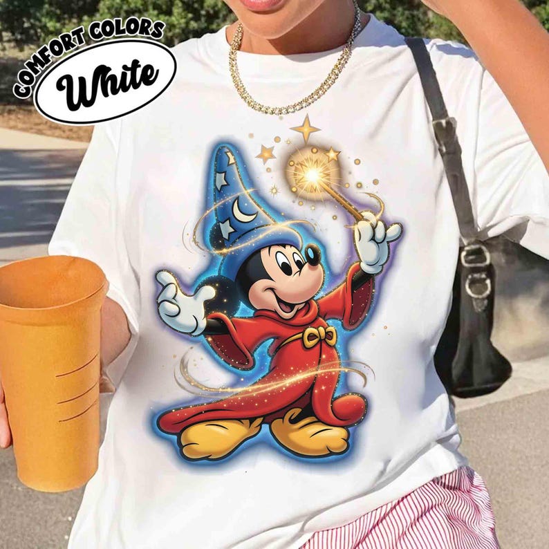 May include: White t-shirt featuring a colorful graphic of Mickey Mouse dressed as a wizard, holding a glowing wand. The design includes a blue wizard hat with stars and moons, and a red robe. The shirt has the words "COMFORT COLORS" and "White" printed on it.