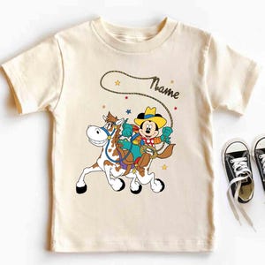 K&ouml;nnte beinhalten: Cremefarbenes T-Shirt mit einem Cartoon-Cowboy, der auf einem Pferd reitet. Der Cowboy tr&auml;gt einen gelben Hut und ein blaues Hemd. Das Pferd ist braun und wei&szlig;. Das Wort "Name" steht &uuml;ber dem Cowboy. Schwarze Turnschuhe mit wei&szlig;en Schn&uuml;rsenkeln liegen neben dem Shirt.