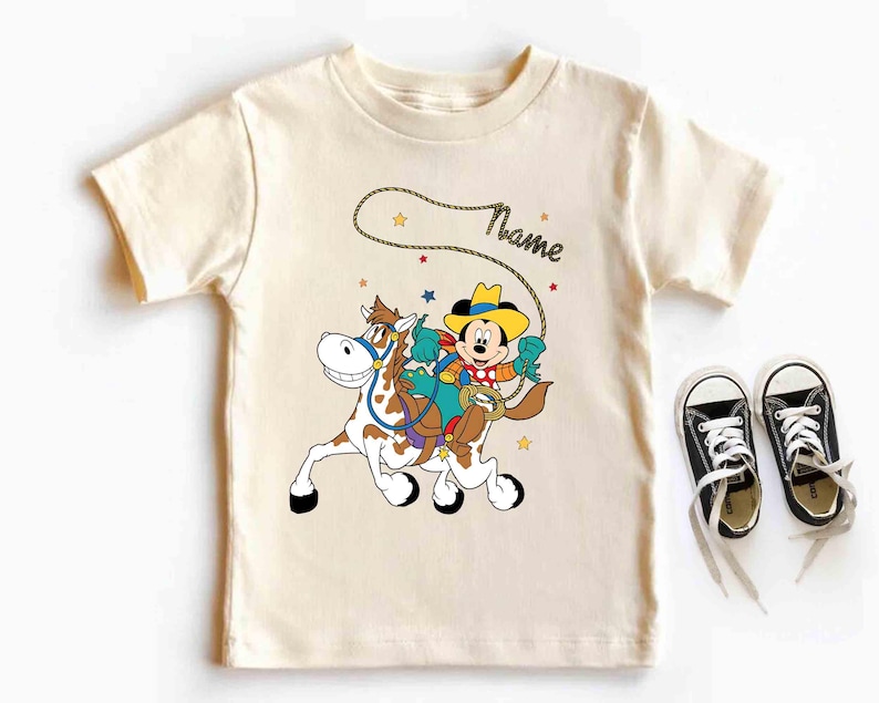 K&ouml;nnte beinhalten: Cremefarbenes T-Shirt mit einem Cartoon-Cowboy, der auf einem Pferd reitet. Der Cowboy tr&auml;gt einen gelben Hut und ein blaues Hemd. Das Pferd ist braun und wei&szlig;. Das Wort "Name" steht &uuml;ber dem Cowboy. Schwarze Turnschuhe mit wei&szlig;en Schn&uuml;rsenkeln liegen neben dem Shirt.