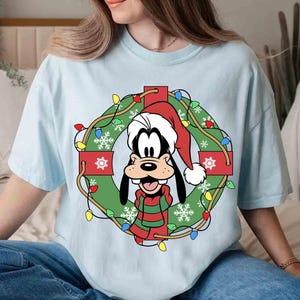 Op de afbeelding: Lichtblauw T-shirt met een cartoonafbeelding van Goofy met een kerstmuts en een groen-rode gestreepte sjaal, in een kerstkrans met lichtjes en sneeuwvlokken. Het shirt heeft een relaxte pasvorm.