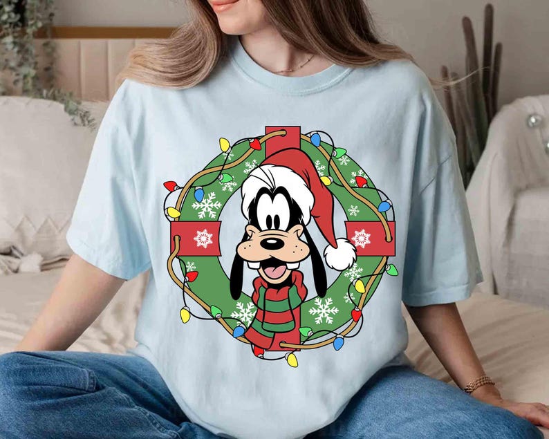 Op de afbeelding: Lichtblauw T-shirt met een cartoonafbeelding van Goofy met een kerstmuts en een groen-rode gestreepte sjaal, in een kerstkrans met lichtjes en sneeuwvlokken. Het shirt heeft een relaxte pasvorm.