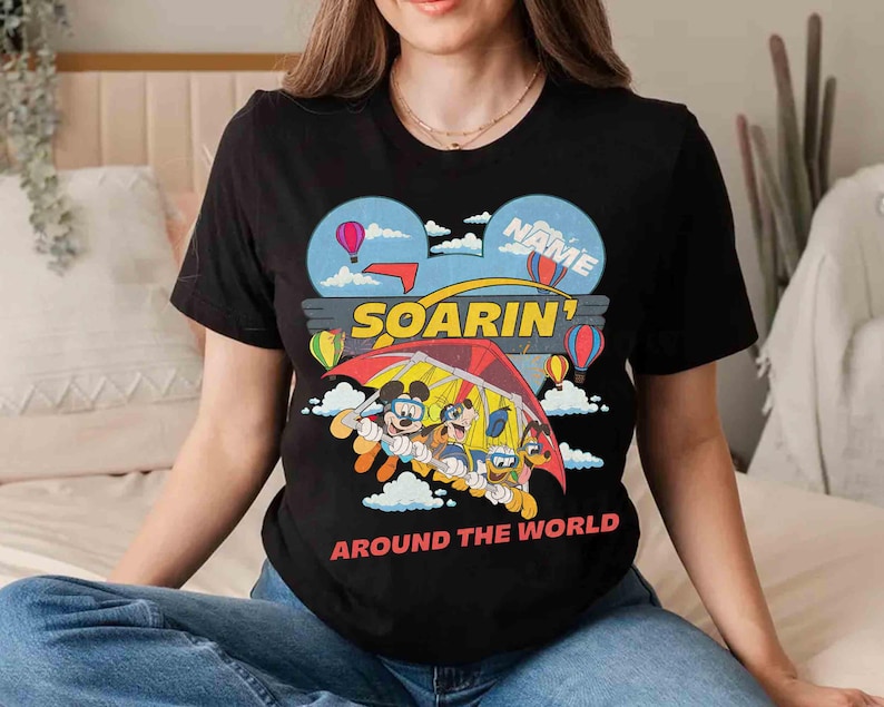 May include: Black t-shirt featuring a colorful graphic of Mickey Mouse, Donald Duck, Goofy, and Pluto in a hang glider. The design includes hot air balloons and the words "SOARIN' AROUND THE WORLD". The word "NAME" is also visible.