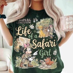 May include: Dark green t-shirt with a graphic featuring Disney's Animal Kingdom theme. The design includes the text "Life of a Safari Girl" with images of Simba, Timon, Pumbaa, and Minnie Mouse in safari attire. The shirt has a crew neck.