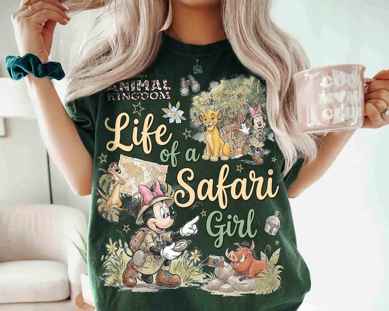 May include: Dark green t-shirt with a graphic featuring Disney's Animal Kingdom theme. The design includes the text "Life of a Safari Girl" with images of Simba, Timon, Pumbaa, and Minnie Mouse in safari attire. The shirt has a crew neck.