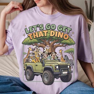 May include: A light purple t-shirt with the text "LET'S GO GET THAT DINO" above a cartoon illustration of a safari vehicle with Mickey Mouse, Donald Duck, and other characters. The vehicle is olive green.