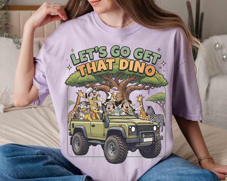 May include: A light purple t-shirt with the text "LET'S GO GET THAT DINO" above a cartoon illustration of a safari vehicle with Mickey Mouse, Donald Duck, and other characters. The vehicle is olive green.
