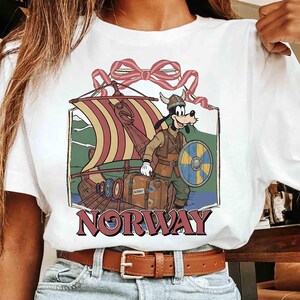 May include: White t-shirt featuring a cartoon character dressed as a Viking, standing next to a longboat. The graphic includes a shield, luggage, and the word "NORWAY" in maroon. A red bow is at the top of the design.
