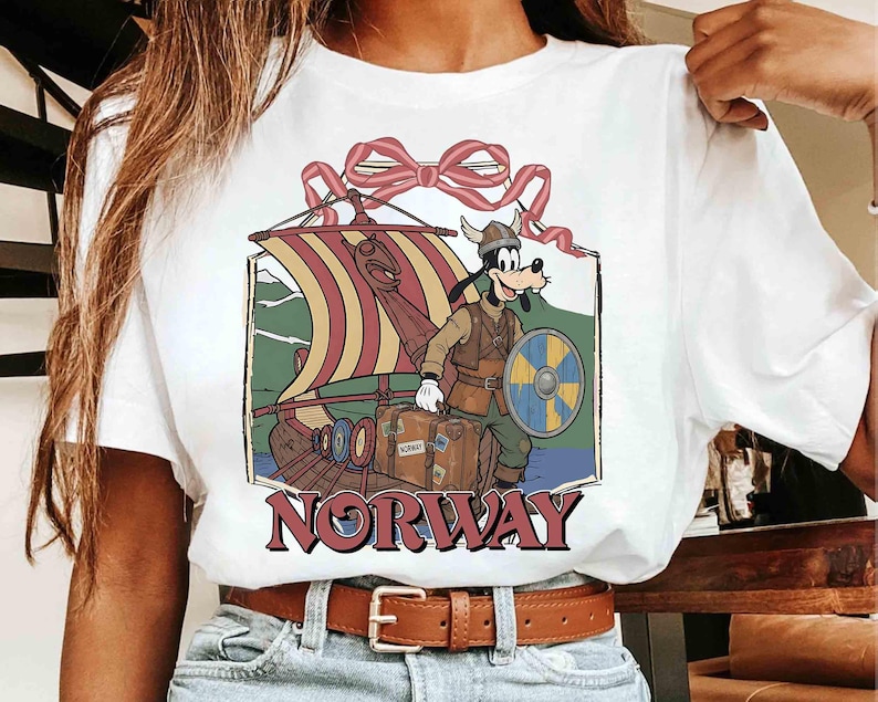 May include: White t-shirt featuring a cartoon character dressed as a Viking, standing next to a longboat. The graphic includes a shield, luggage, and the word "NORWAY" in maroon. A red bow is at the top of the design.