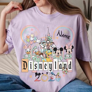 May include: A light purple t-shirt featuring a Disneyland graphic with Mickey Mouse, Minnie Mouse, Donald Duck, and other Disney characters. The shirt includes the text "Disneyland 2020" and a space for a name.