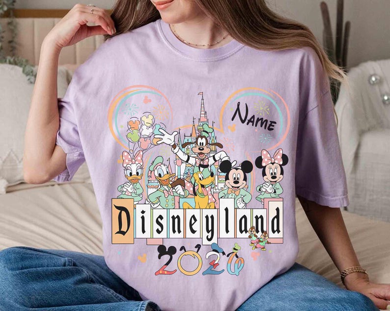 May include: A light purple t-shirt featuring a Disneyland graphic with Mickey Mouse, Minnie Mouse, Donald Duck, and other Disney characters. The shirt includes the text "Disneyland 2020" and a space for a name.