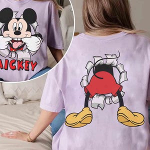 May include: A light purple t-shirt featuring a graphic of Mickey Mouse bursting through a torn paper effect. The front shows Mickey's face with the word "Mickey" in red. The back shows Mickey's legs, red shorts, and yellow shoes.