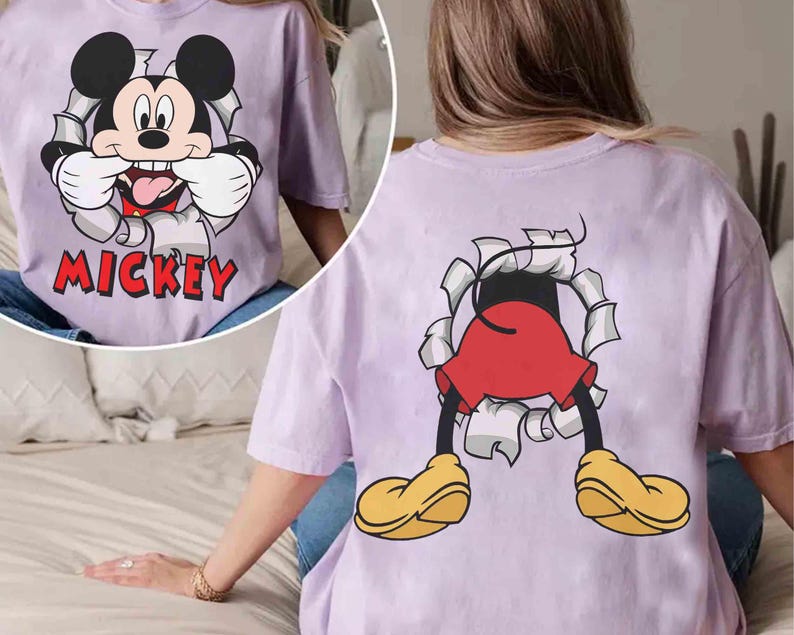 May include: A light purple t-shirt featuring a graphic of Mickey Mouse bursting through a torn paper effect. The front shows Mickey's face with the word "Mickey" in red. The back shows Mickey's legs, red shorts, and yellow shoes.