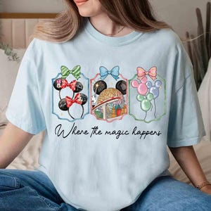 May include: Light blue t-shirt featuring three framed illustrations with Minnie Mouse ears, a theme park scene, and balloons. Each illustration is topped with a colorful bow. The text "Where the magic happens" is written below the illustrations.