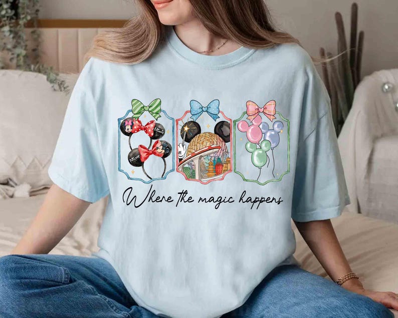 May include: Light blue t-shirt featuring three framed illustrations with Minnie Mouse ears, a theme park scene, and balloons. Each illustration is topped with a colorful bow. The text "Where the magic happens" is written below the illustrations.
