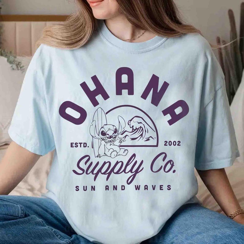 May include: Light blue t-shirt with a purple graphic featuring the word "OHANA" above a cartoon character with a surfboard and a wave. The text "Supply Co." and "Sun and Waves" are also included.