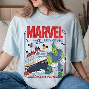 May include: Light blue t-shirt featuring a graphic with the text "MARVEL Day at Sea". The design includes a cruise ship, cartoon characters, and the option for a custom name. The shirt is a light blue color.