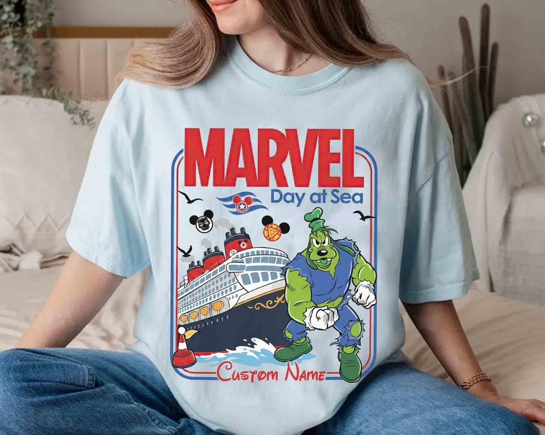 May include: Light blue t-shirt featuring a graphic with the text "MARVEL Day at Sea". The design includes a cruise ship, cartoon characters, and the option for a custom name. The shirt is a light blue color.