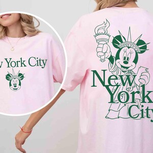 May include: A light pink t-shirt with the text "New York City" in green. The shirt features a graphic of Minnie Mouse as the Statue of Liberty. The same design is on the front and back of the shirt.