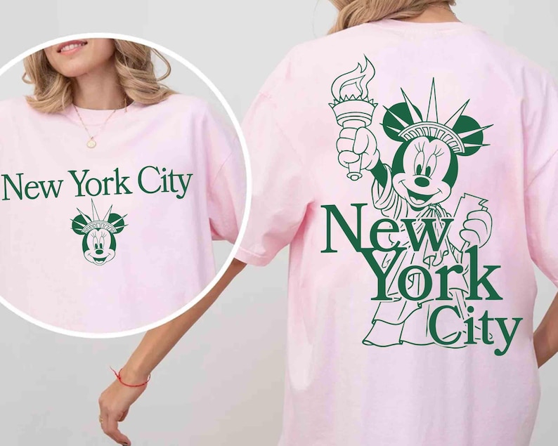 May include: A light pink t-shirt with the text "New York City" in green. The shirt features a graphic of Minnie Mouse as the Statue of Liberty. The same design is on the front and back of the shirt.
