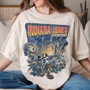 May include: A white t-shirt with a graphic print of Mickey Mouse dressed as Indiana Jones. The text "Indiana Jones Adventure" is printed above Mickey Mouse.