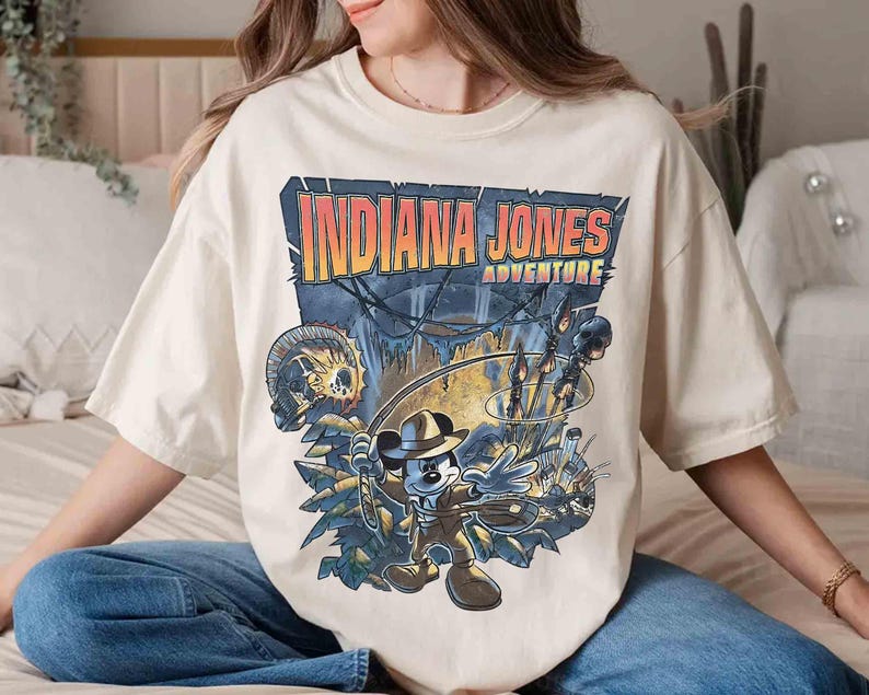 May include: A white t-shirt with a graphic print of Mickey Mouse dressed as Indiana Jones. The text "Indiana Jones Adventure" is printed above Mickey Mouse.