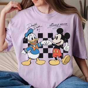 May include: A light purple t-shirt featuring Donald Duck and Mickey Mouse. Donald Duck is wearing a blue sailor suit and Mickey Mouse is wearing red shorts. The text "Donald Duck #1" and "Mickey Mouse" are above the characters.